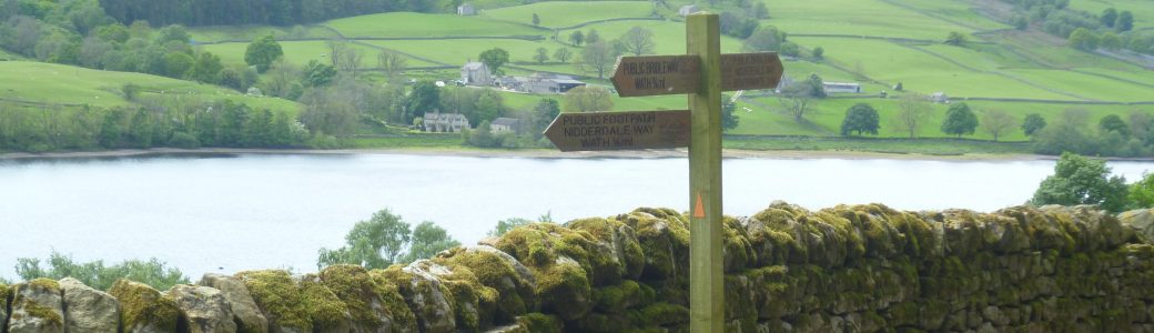 A wooden Nidderdale Way footpath sign with a dry stone wall, reservoir and the valley side beyond
